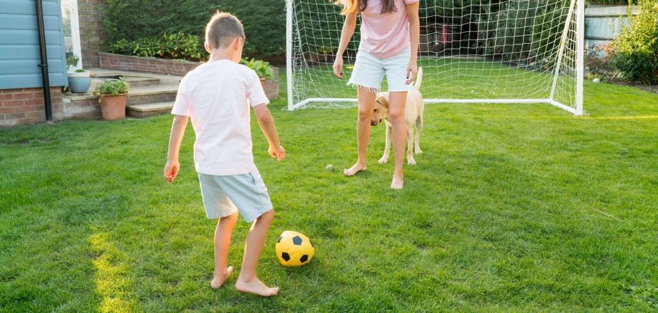 A mother playing soccer with her son in the back garden with a dog and soccer net behind them