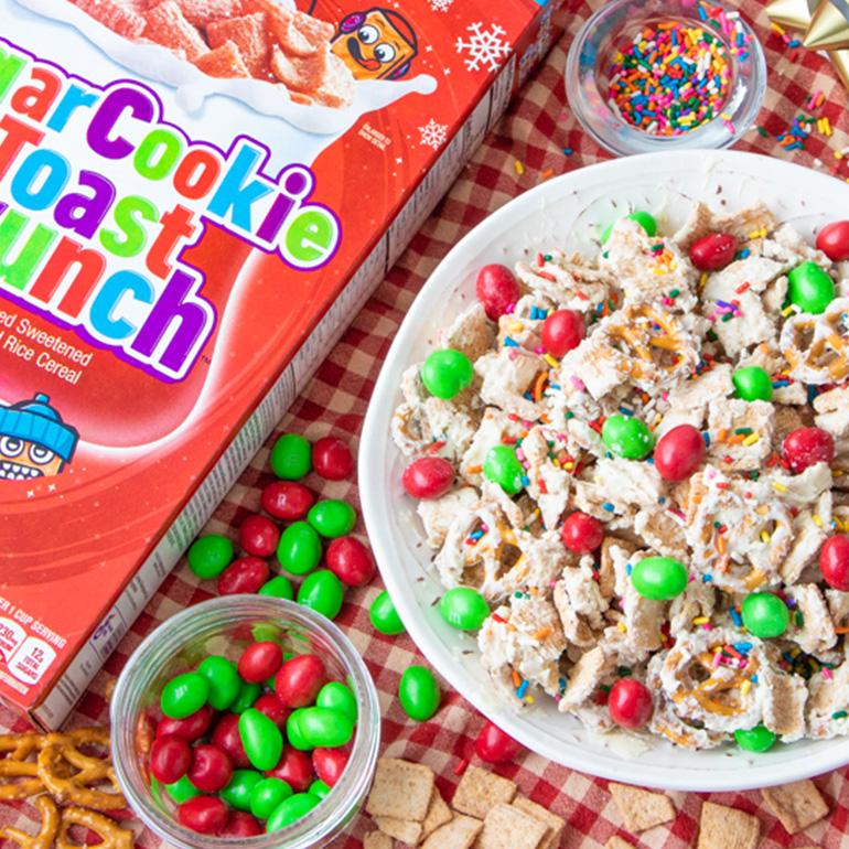 Una caja de Sugar Cookie Toast Crunch junto a un bol de aperitivos hechos con cereales. Al lado hay dos tazones pequeños llenos de chocolate y chispas de colores.