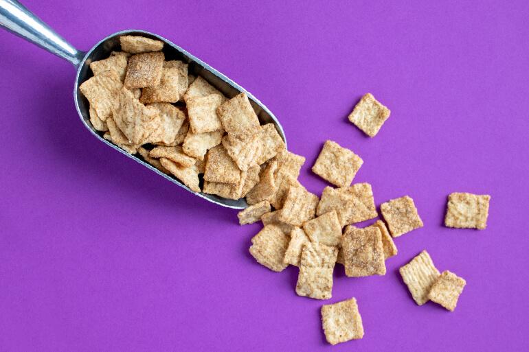 Una pala metálica de mano llena de Cinnamon Toast Crunch. Parte de los cereales se han derramado de la pala y están esparcidos por la superficie.