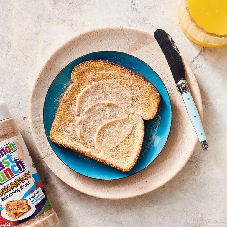 Buttered Toast with Cinnadust™ on a plate with a butter knife, a glass of orange juice and a jar of Cinnadust™ beside it.